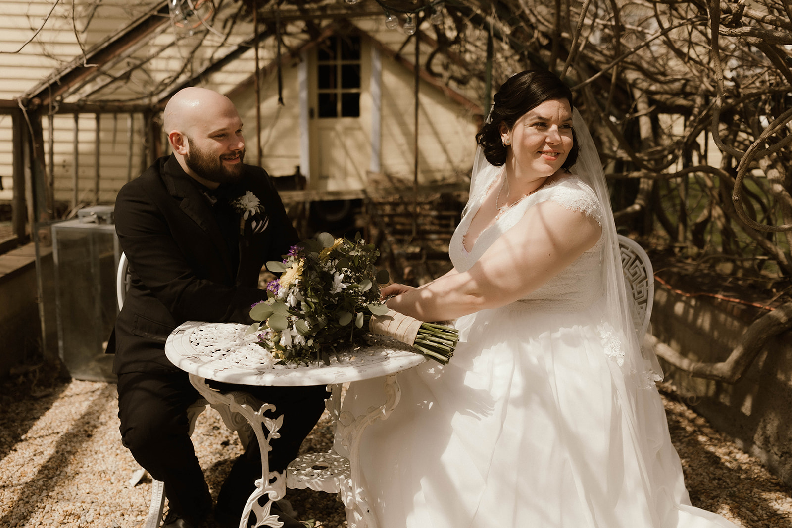 Beautiful bride and groom sit at small outdoor table and pose together