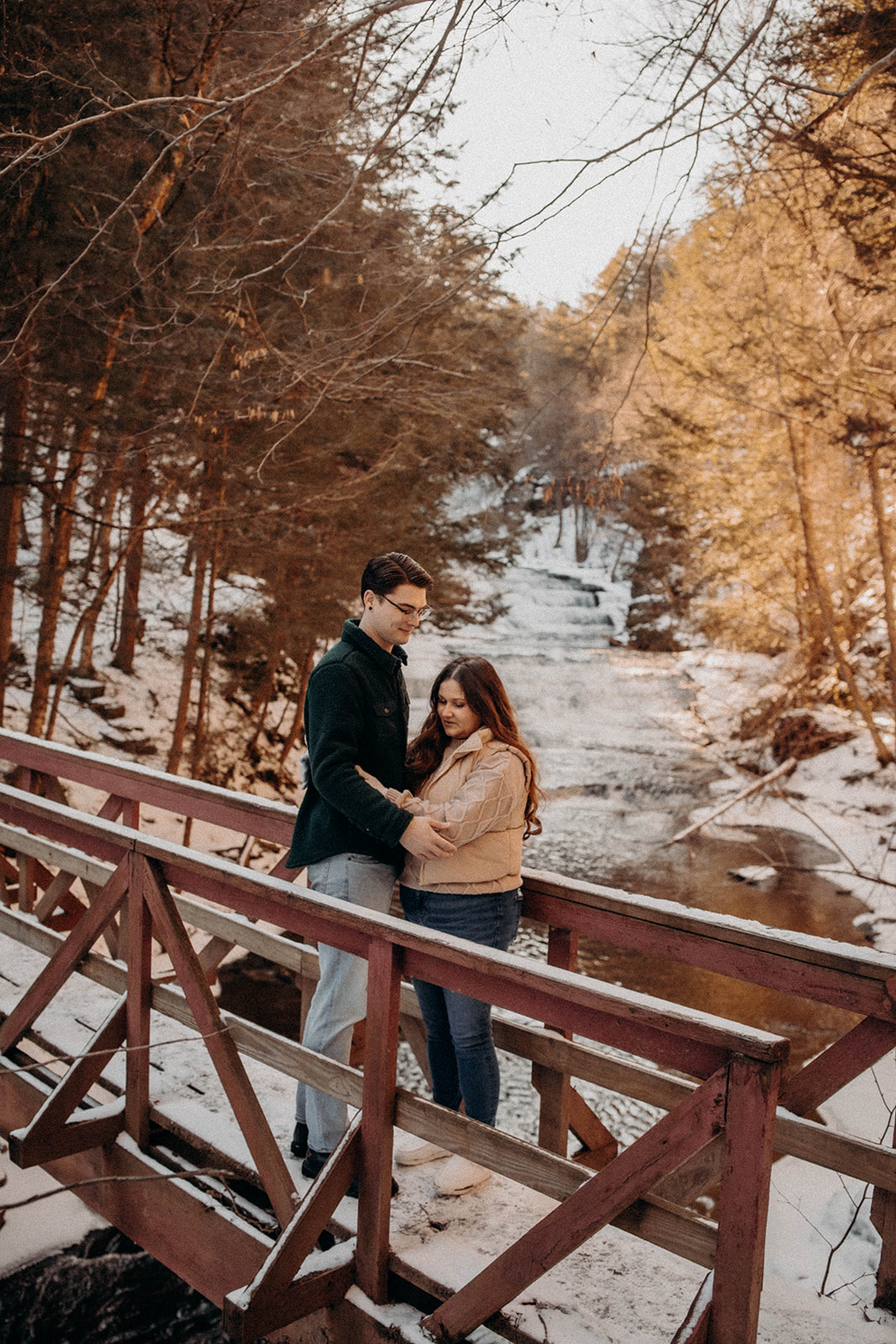 Beautiful couple stop for a moment on a striking red bridge in the snow during their winter engagement photos at Huyck preserve