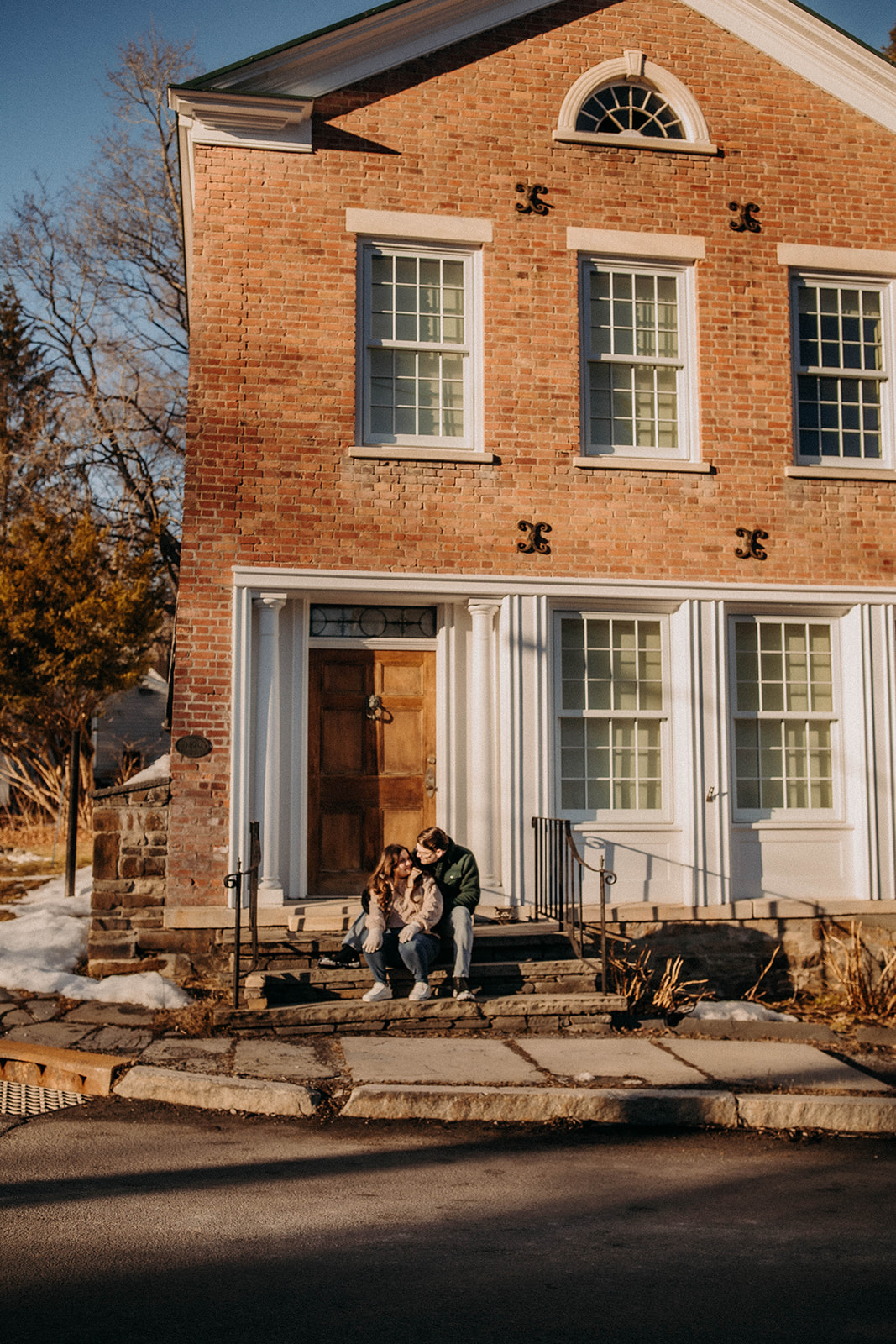 Couple sits outside a historic building during their winter engagement photoshoot in Rensselaerville New York!
